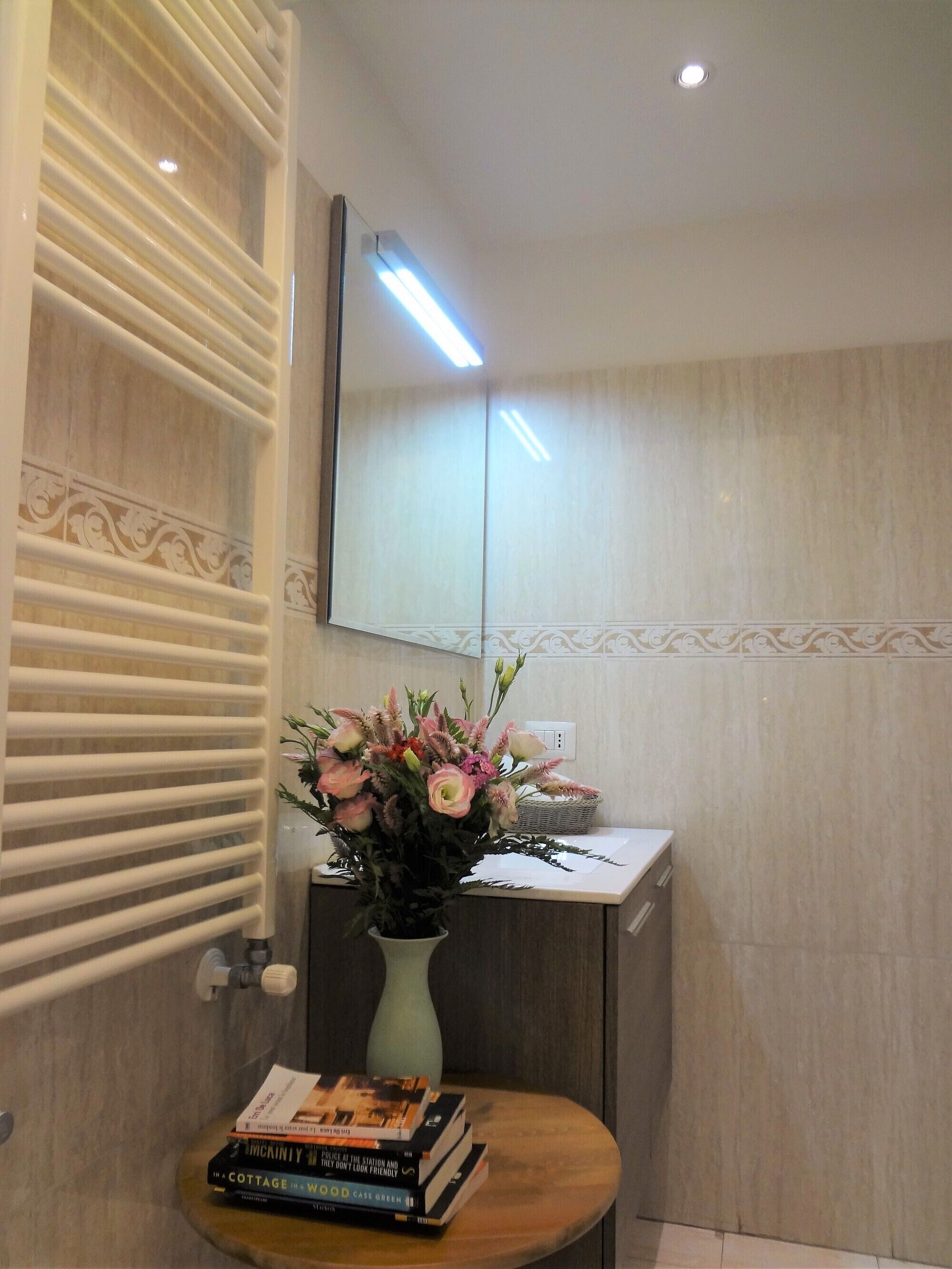 Ein Badezimmer mit einem Spiegel, einem Handtuchhalter, einer Vase mit Blumen und einem Stapel Bücher auf einem kleinen Tisch.