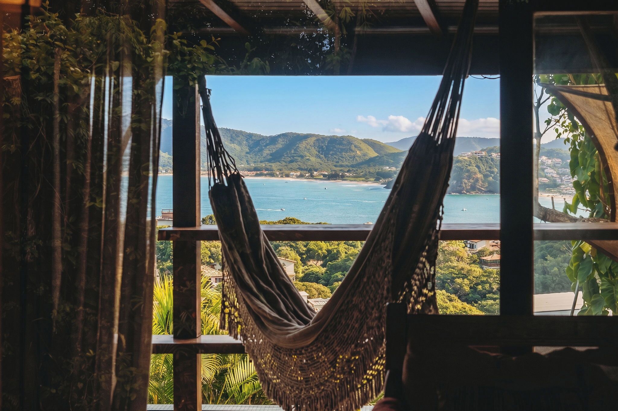 Una hamaca colgada junto a una ventana con vistas a un paisaje costero con colinas y una playa.
