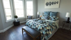 Ein Schlafzimmer mit einem Bett, einer Bank, zwei Fenstern, einer Lampe und einem Wandbild.
