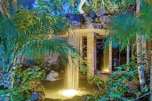 Een waterval in een weelderige tuin, met een stenen muur en een glazen deur die naar een kamer leidt.