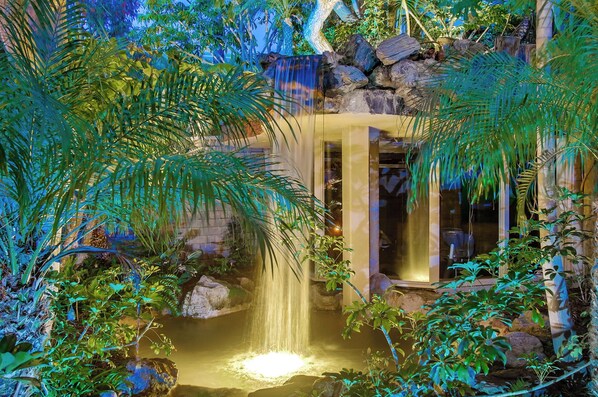 A waterfall in a lush garden with a stone wall and a glass door leading to a room. - North Coast A101 Corner condo West & South Facing Ocean views from Two Balconies (Oceanside)