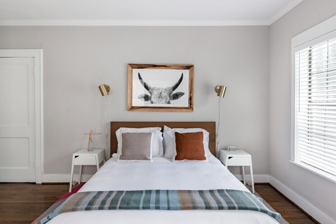 A bedroom with a large bed, two bedside tables, a framed picture on the wall, and a window with blinds.