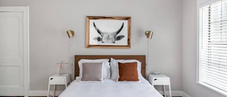 A bedroom with a large bed, two bedside tables, a framed picture on the wall, and a window with blinds.