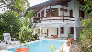 Una casa de dos pisos con piscina, rodeada de abundante vegetación.