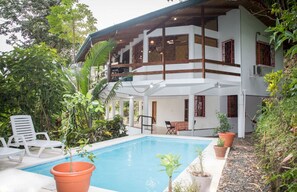 A two-story house with a swimming pool, surrounded by lush greenery.