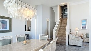 En rummelig spiseplads med et stort marmorbord, hvide polstrede stole, en moderne lysekrone og en trappe til et niveau oppe.