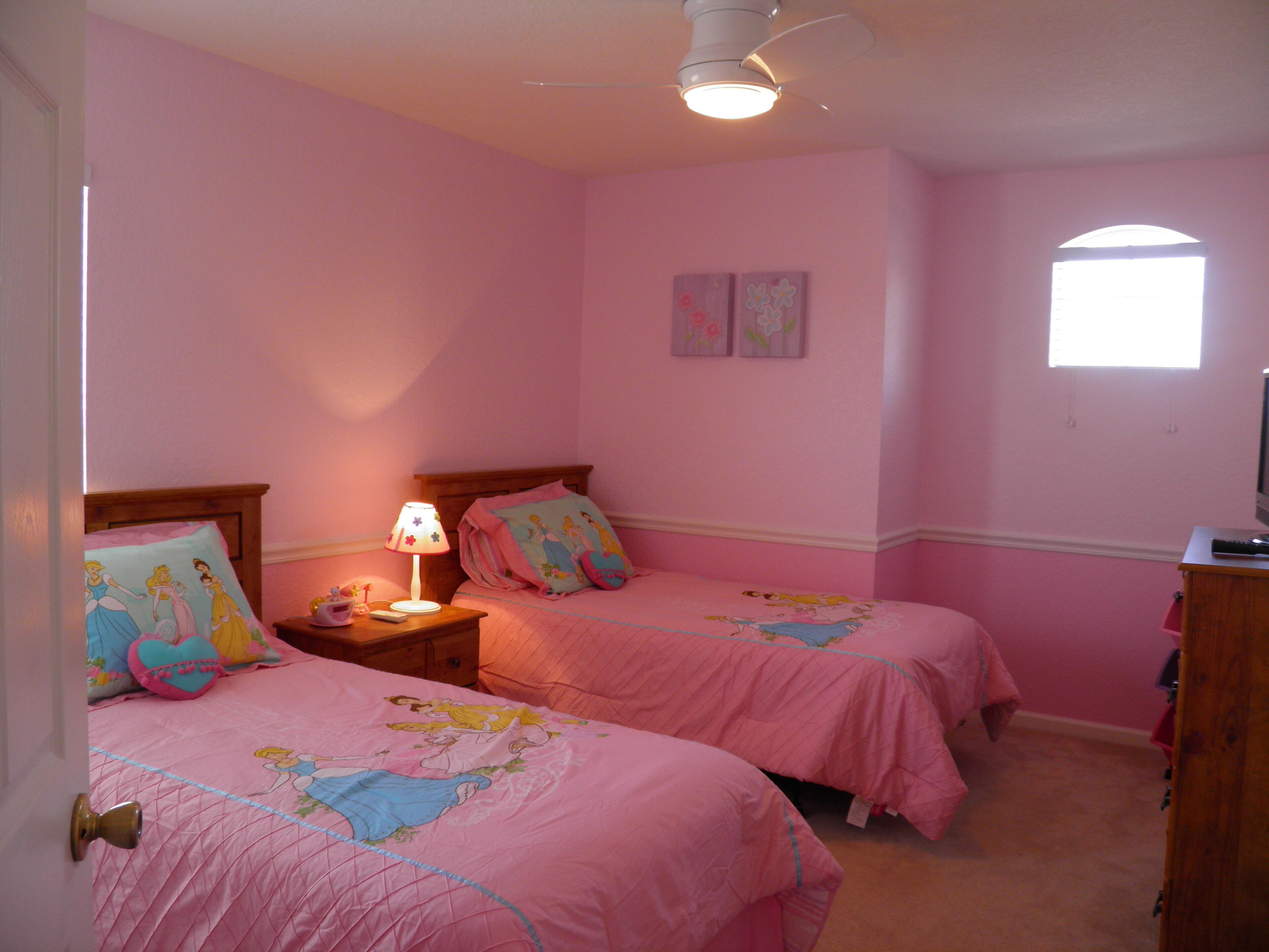 A pink-themed bedroom with two beds, a nightstand, a lamp, and a ceiling fan.