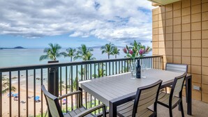 A balcony with a wooden table and chairs, overlooking a beach with palm trees and a view of the ocean.
