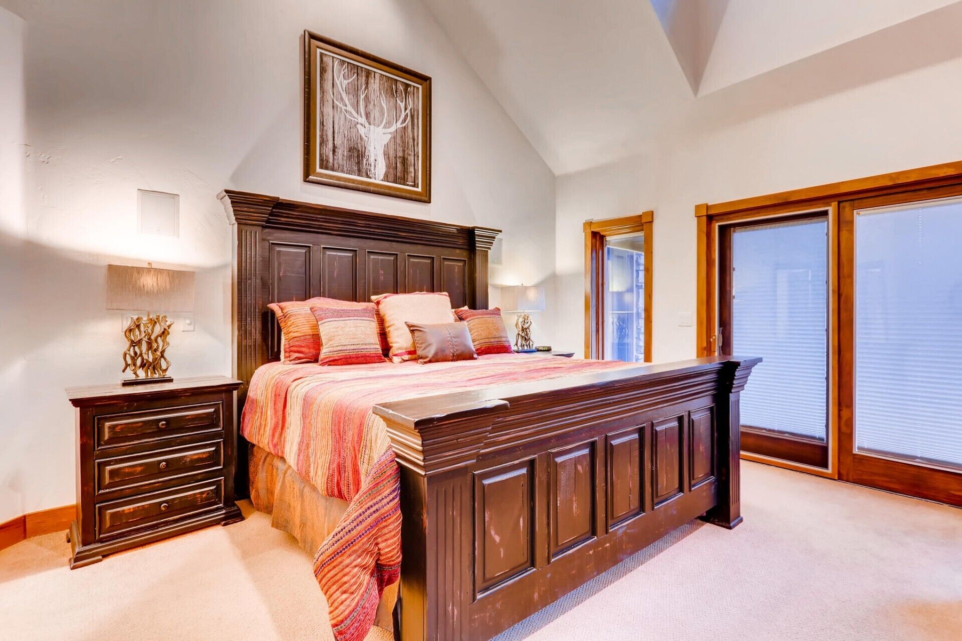 Une chambre à coucher avec un grand lit, une tête de lit en bois, une table de chevet, une lampe et un tableau représentant une tête de cerf encadrée.