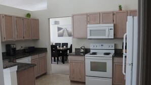 A kitchen with wooden cabinets, a microwave, an oven, and a refrigerator. There is a dining area with chairs and a table. - Beautiful Waterside Villa For Rent In Orlando (Clermont)