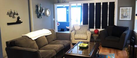 A living room with a brown sofa, a beige armchair, and a glass coffee table. There is a ceiling fan, a window with curtains, and a rug with a leaf pattern.