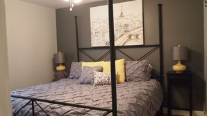 Ett sovrum med en säng, två sängbord, en takfläkt och en stor målning med ett stadsmotiv på väggen.
