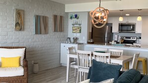 A modern kitchen with white cabinets, a stainless steel refrigerator, and a microwave. There is a dining area with white chairs and a table.