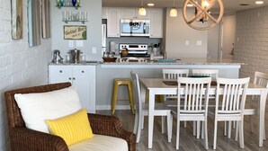 A modern kitchen with white cabinets, a central island, and a dining area with white chairs and a wicker sofa with a yellow pillow.