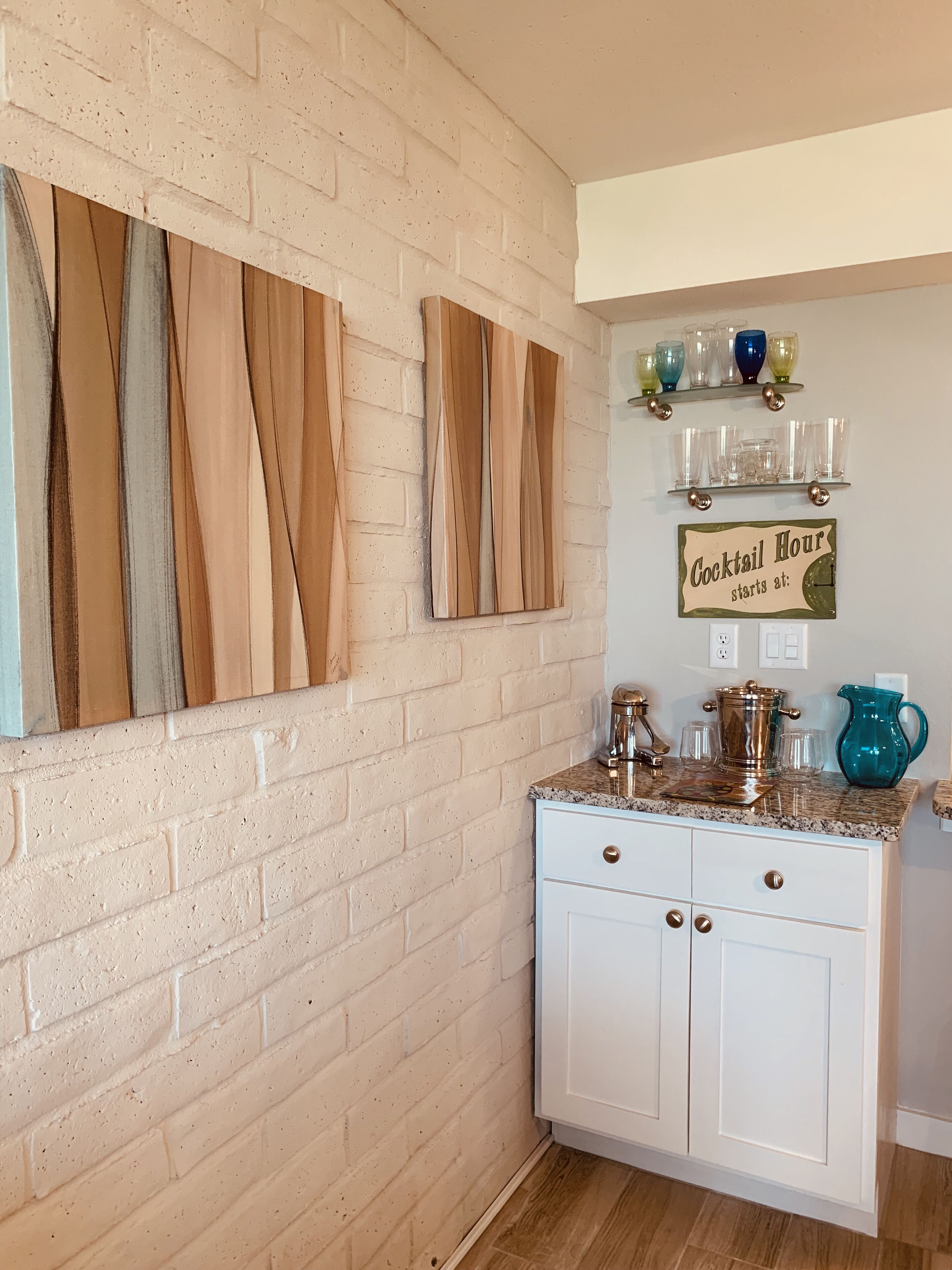 Una cocina con armarios blancos, encimera de granito y pared de ladrillo. Hay listones de madera en la pared, un estante con cristalería y un letrero que indica 'Cocktail Hour starts at:'.