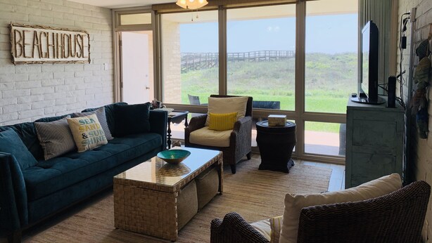 A living room with a large window, a ceiling fan, a sign reading "BEACHHOUSE", and a view of a bridge and greenery outside.