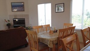 Un coin repas avec une table dressée pour six personnes, entourée de chaises en bois. On aperçoit un canapé marron au fond et un espace cuisine avec un réfrigérateur et une cuisinière.