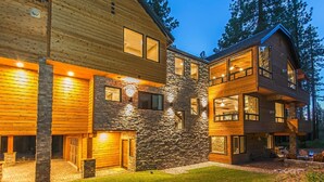 A two-story log cabin with a stone facade and large windows, surrounded by a well-maintained lawn and tall trees.