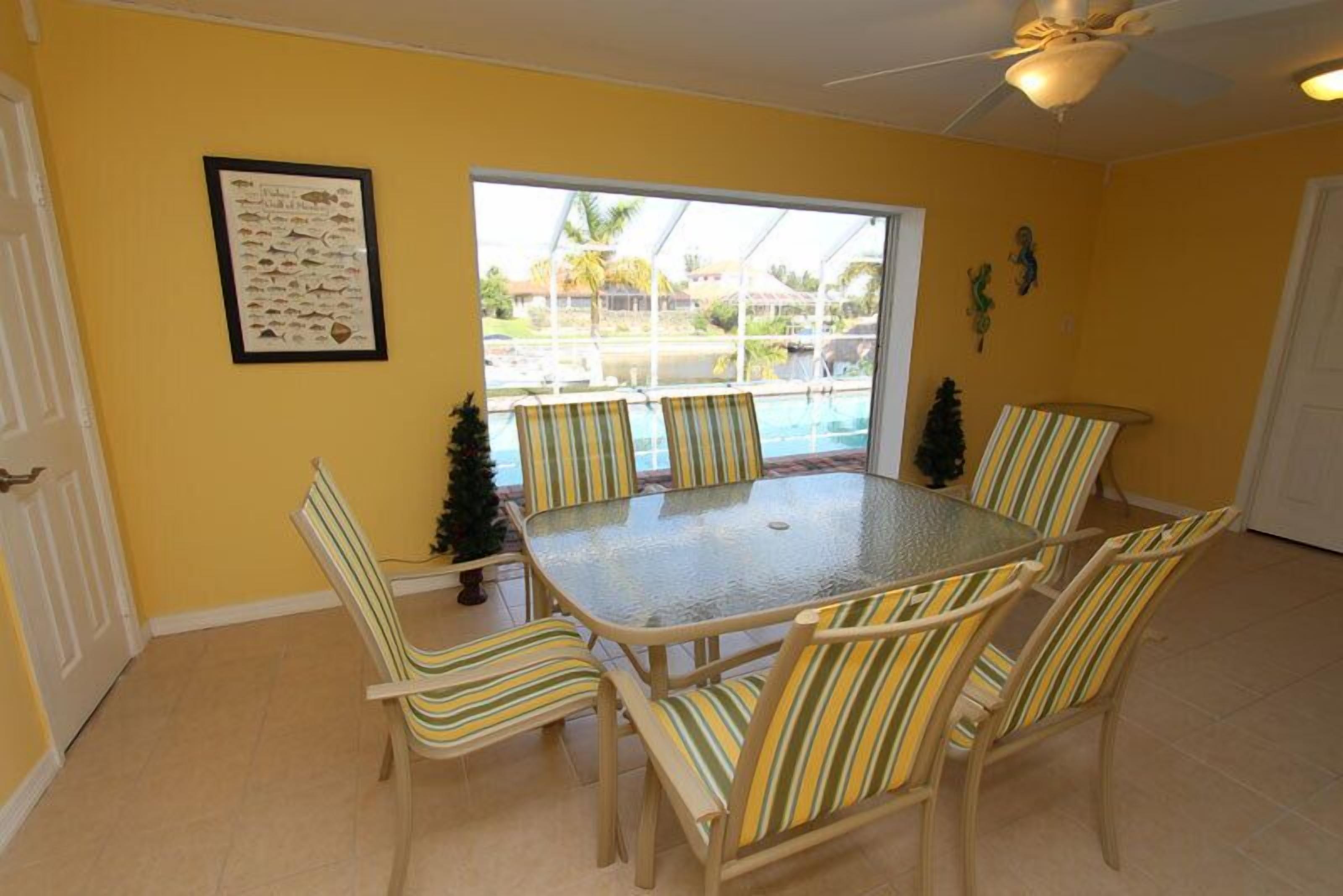 Un comedor con mesa de vidrio y sillas a rayas, un cartel enmarcado con un pez en la pared, y vistas a una piscina y palmeras a través de un ventanal grande.
