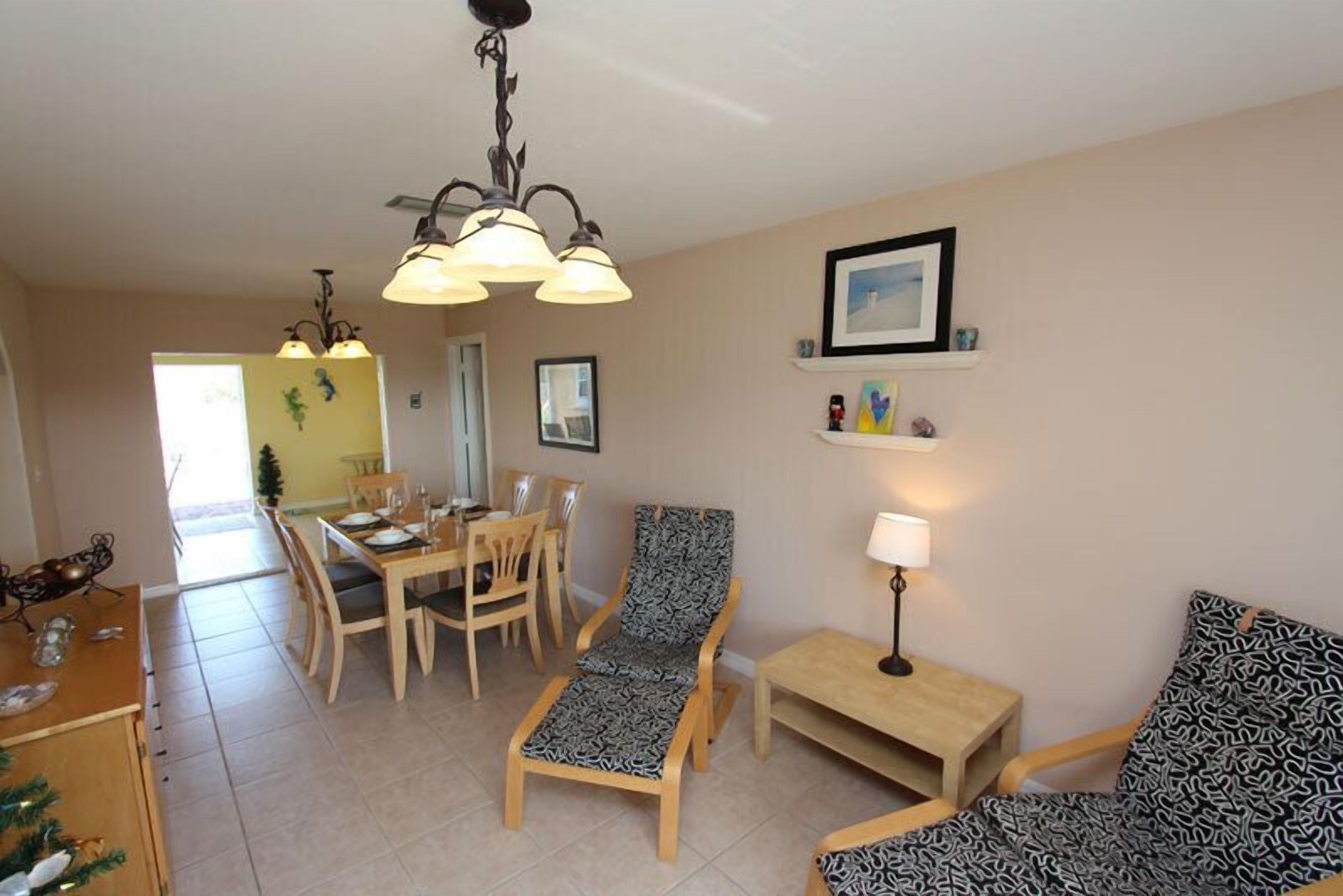 A dining area with a table and chairs, a lounge chair, a small table with a lamp, and a wall-mounted shelf with pictures.