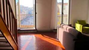 Ein Wohnzimmer mit einer Couch, einem grünen Sessel, einer Holztreppe und Blick auf die Stadt durch ein großes Fenster.