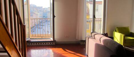 A living room with a sofa, a green chair, a wooden staircase, and a view of the city through a large window.