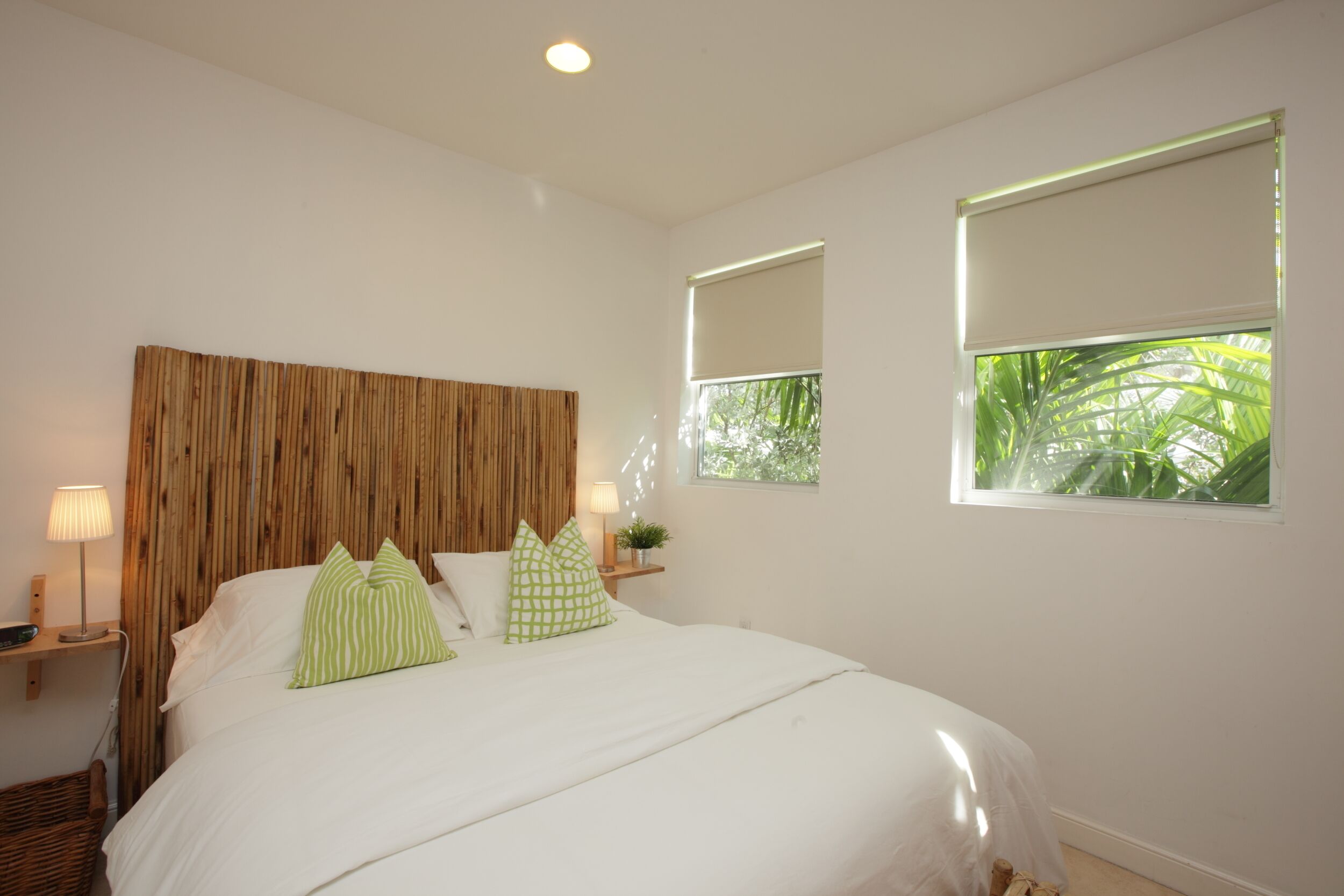 A bedroom with a wooden headboard, a bed with white bedding and green pillows, two bedside tables with lamps, and windows with blinds.
