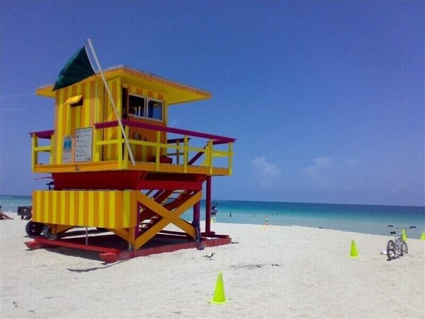 A lifeguard tower with a red and yellow striped design, located on a sandy beach with clear blue water in the background.