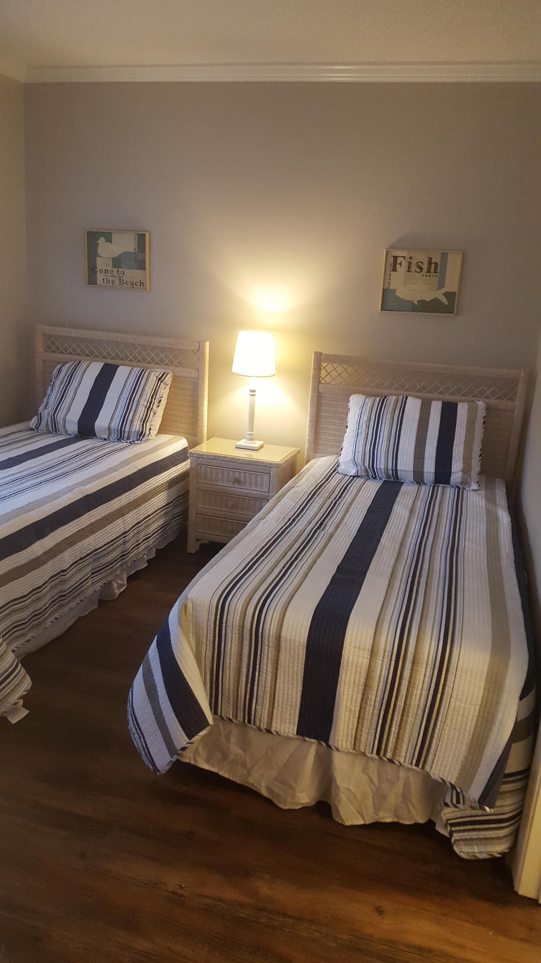 Ein Zimmer mit zwei Betten, jeweils mit gestreifter Bettwäsche und einem Kopfteil. Dazwischen befindet sich ein Nachttisch mit einer Lampe.