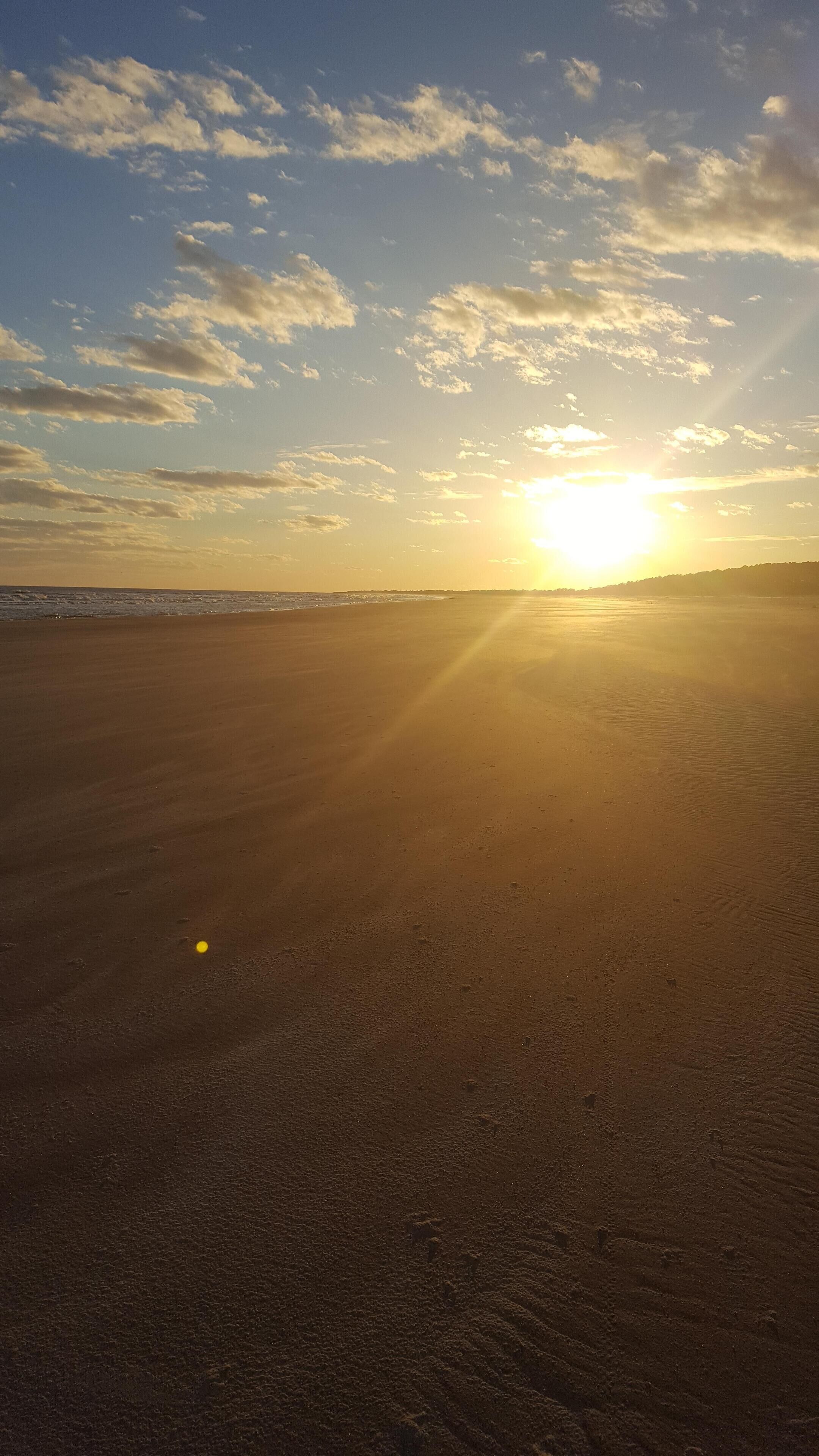 Ein Strand bei Sonnenuntergang, die Sonne tief am Horizont, wirft ein warmes Licht über den Sand.