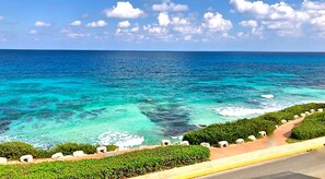A coastal view with clear turquoise waters, a paved walkway, and lush greenery.
