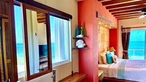 Ein Hotelzimmer mit großem Fenster, einem hölzernen Schminktisch mit Waschbecken und einem Bett mit Meerblick, sowie einer roten Akzentwand.