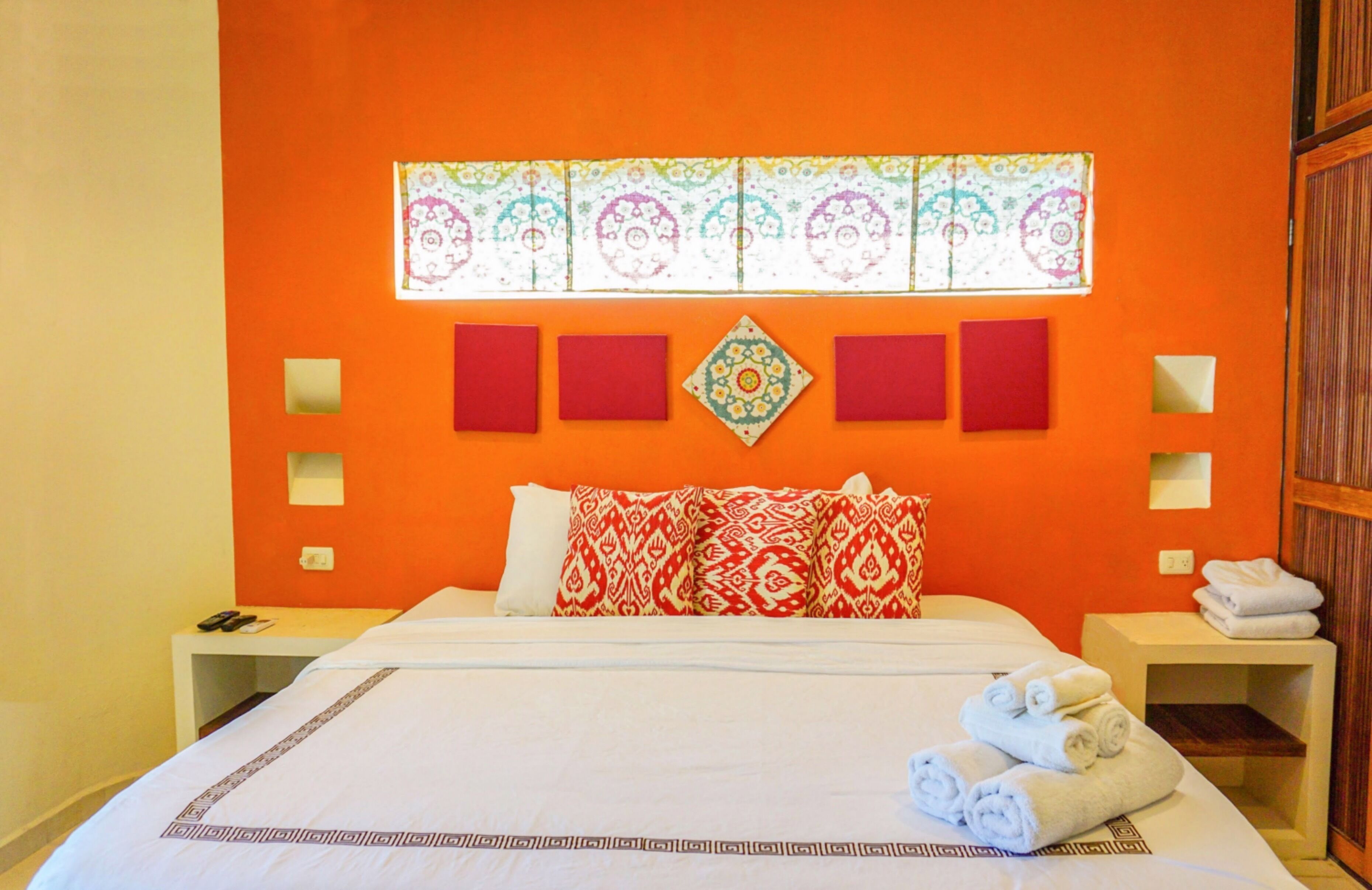 Una habitación de hotel con una cama grande, almohadas con estampado rojo y blanco, un adorno en la pared y toallas dobladas con cuidado al lado.