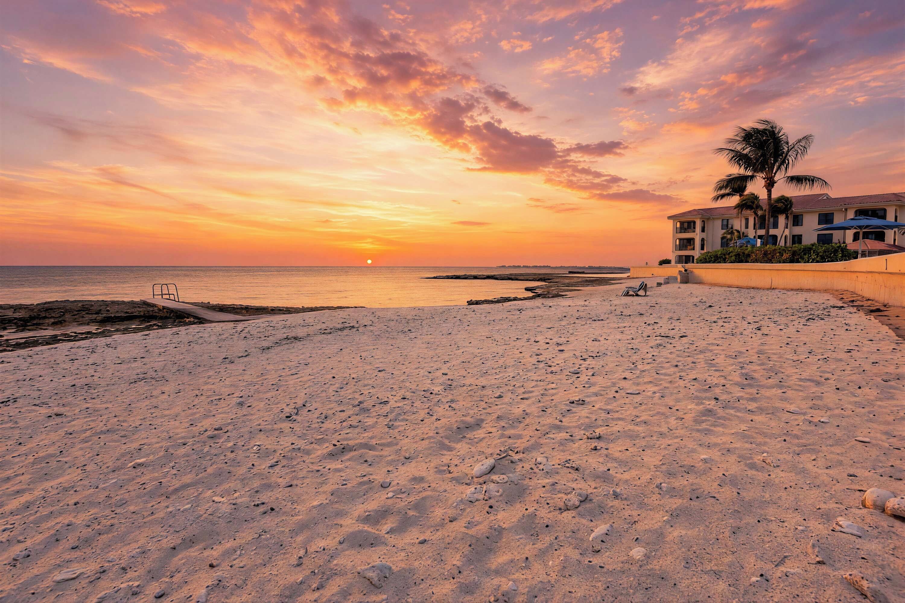 Une plage au coucher du soleil, un bâtiment doté d’un balcon et des palmiers.