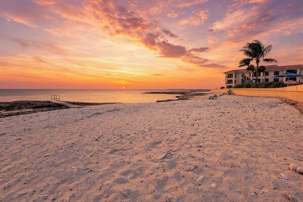 Une plage au coucher du soleil, un bâtiment doté d’un balcon et des palmiers.