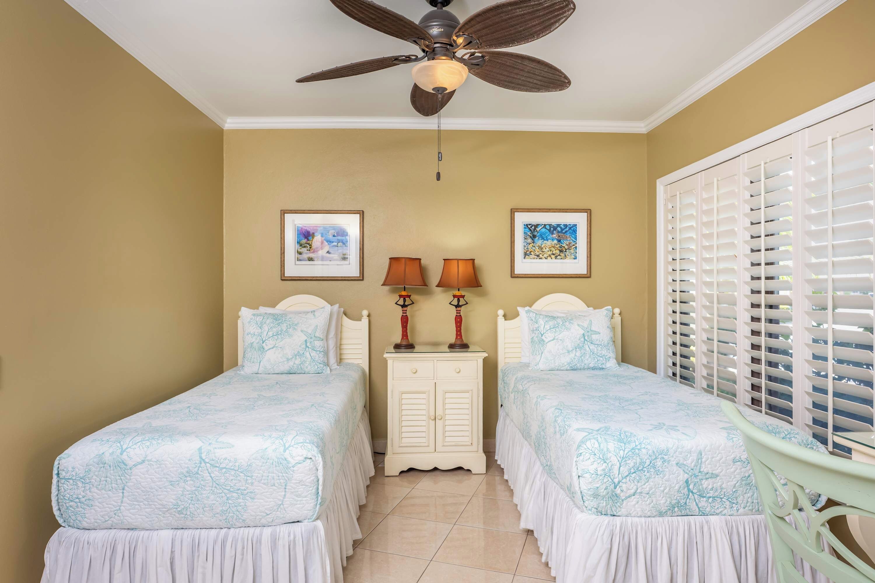 A bedroom with two beds, a ceiling fan, a nightstand with lamps, and a window with blinds.