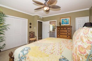 Un dormitorio con cama, ventilador de techo, cómoda, una silla y una puerta que da a un baño.