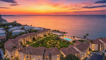 A resort with multiple buildings, a swimming pool, and a view of the ocean at sunset.