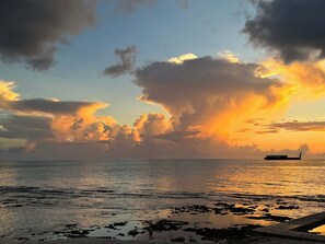 A sunset over a calm sea with a ship in the distance and a rocky shoreline.