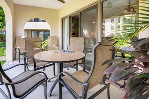 Un patio al aire libre con una mesa redonda y sillas, una puerta de cristal que da a un vestíbulo cubierto, y una vista de un edificio al fondo.