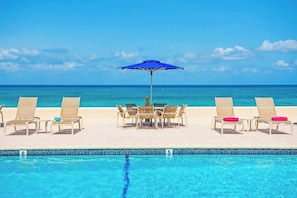 A poolside area with lounge chairs and a table under a blue umbrella, overlooking a clear blue ocean.