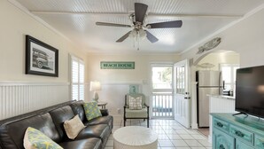 A living room with a black leather couch, a round ottoman, a ceiling fan, a framed picture, a sign reading 'BEACH HOUSE', and a kitchen area visible through an open door. - DownStairs with Great back yard for kiddos (Folly Beach)