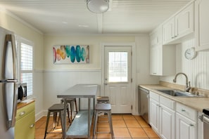 Una cocina con encimera blanca, pileta de acero inoxidable y un microondas. Se observa una puerta con ventana, un cuadro en la pared y un reloj sobre la pileta.
