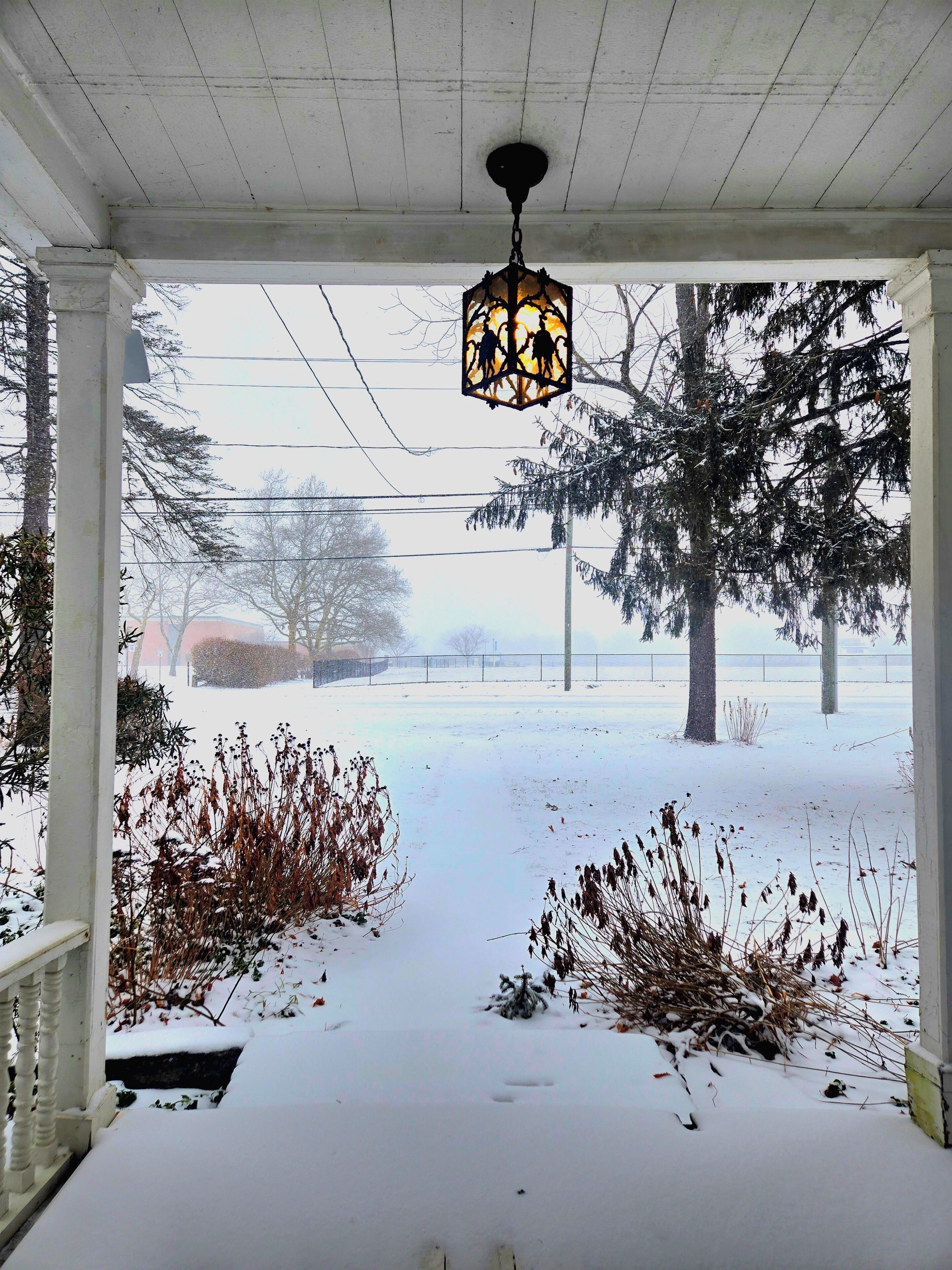 A covered porch with a hanging lantern, snow-covered ground, and a view of a snowy landscape with trees.