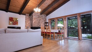 A spacious living area with a sofa, wooden dining table, and chairs. A fireplace is visible in the background, and there is a large window providing a view of the outdoors.