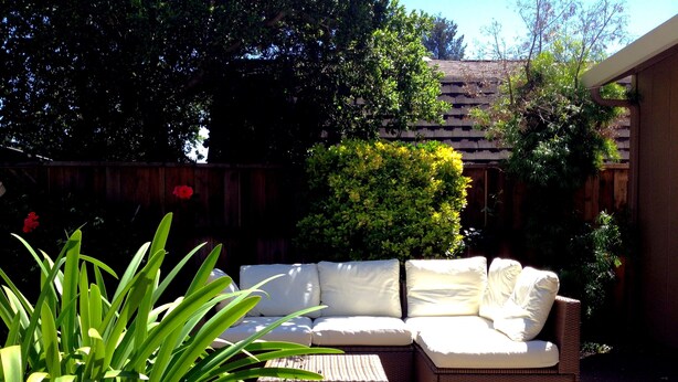 A patio with a wicker sofa and a small table, surrounded by various plants and a wooden fence.