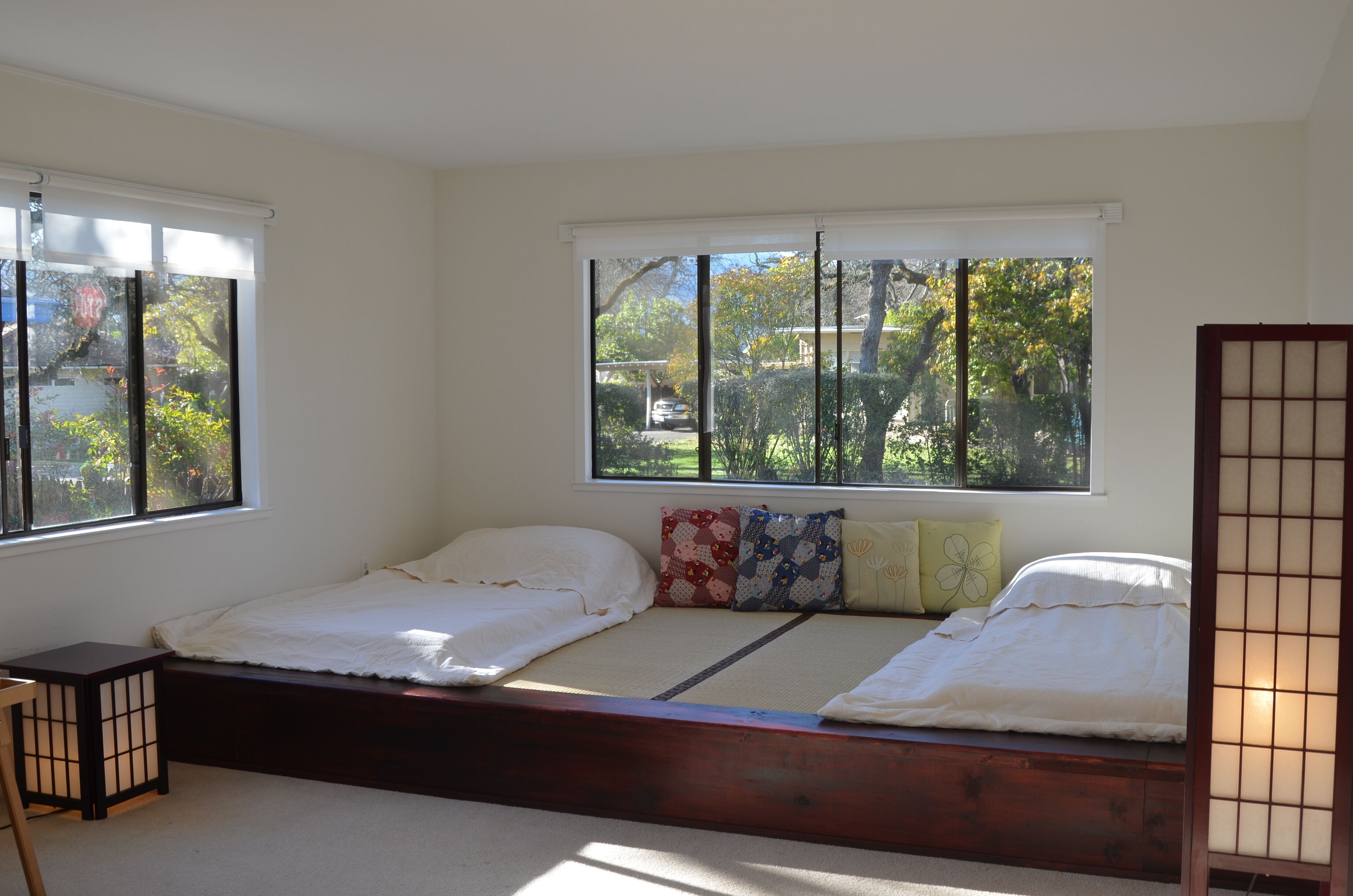 A room with a low wooden bed, a small table, and a window with a view of trees and a car outside.