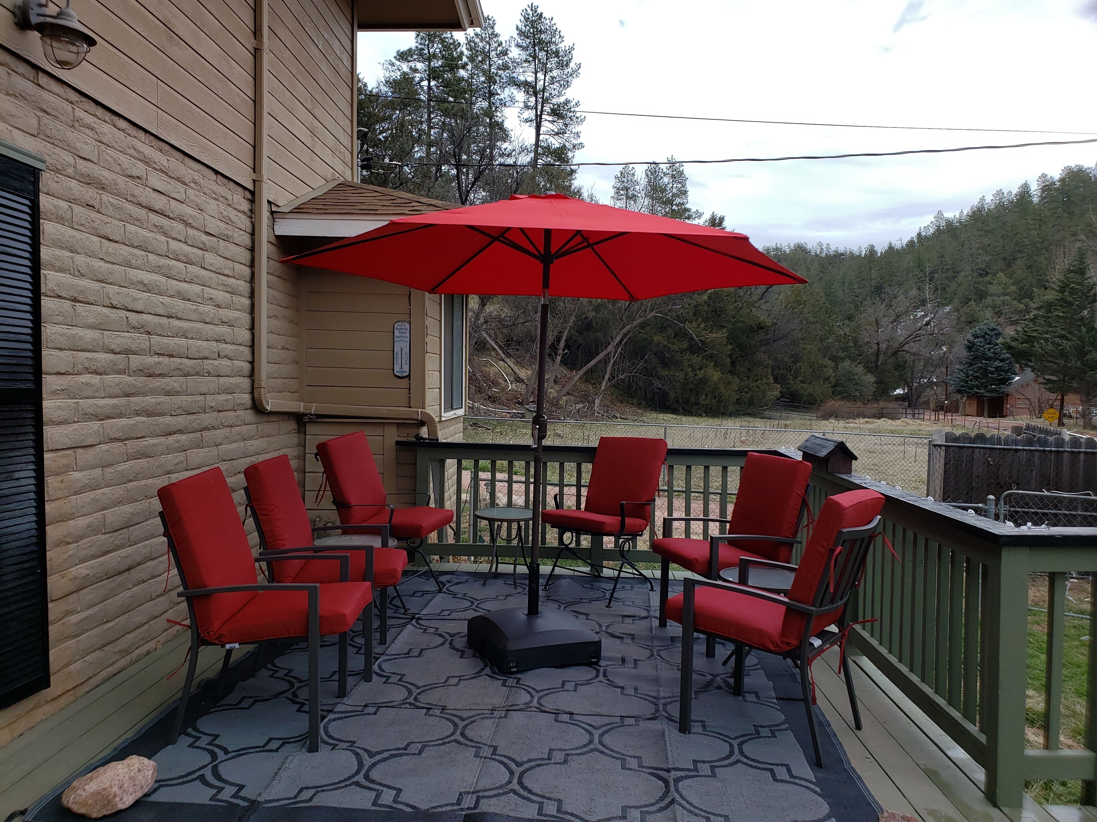 Um alpendre coberto com cadeiras vermelhas e uma mesa, um guarda-sol vermelho, e vista para árvores e uma cabana ao fundo.
