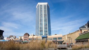 A tall, modern building with a glass facade, surrounded by various commercial buildings and a pedestrian walkway. - DIRECT Oceanfront on Boardwalk — Thanksgiving — Nov 23-27 — 2, 3 or 4 nights (Atlantic City)