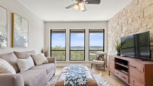 A modern living room with a sofa, ottoman, and entertainment unit, large windows with a view, and a ceiling fan. - Lake Travis Oasis, Pool, Hot Tub, Lake Views, Near Marinas & Austin! (Austin)